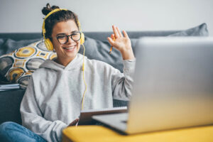 woman with glasses waves to laptop screen while on telehealth therapy